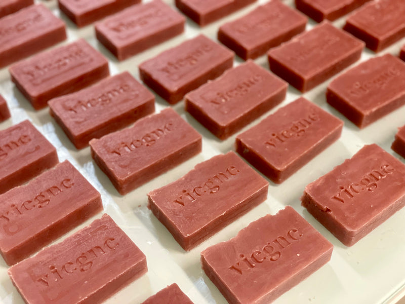 Fabrication artisanale de savons cépages pinot noir ocre rouge ocre rose. Les savons sont alignés sur la table pour être tamponné au logo de la marque Vie.gne.